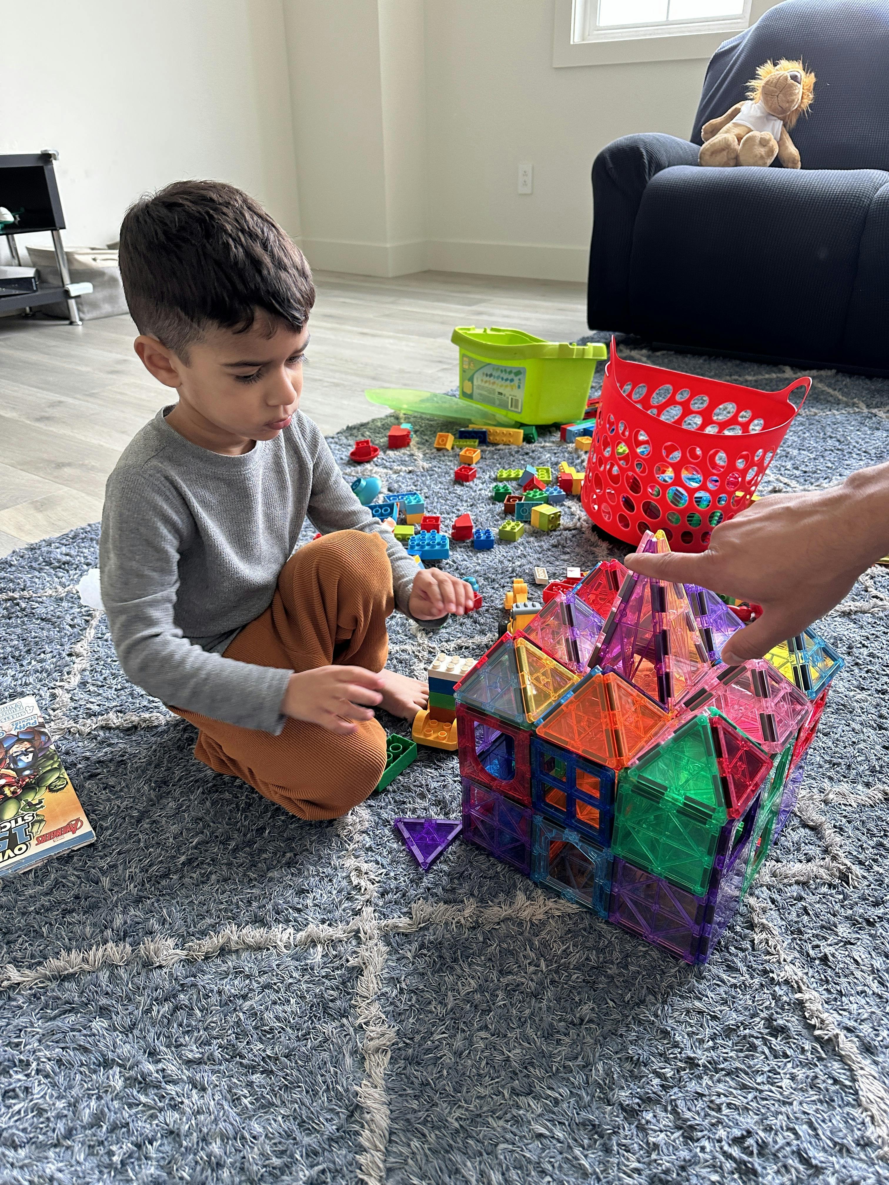 Child building a house with colorful translucent magnetic tiles, person's hand pointing