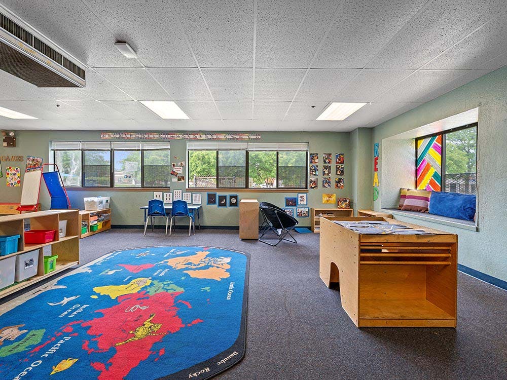 Classroom with world map rug, shelves of toys, reading nook, and small tables