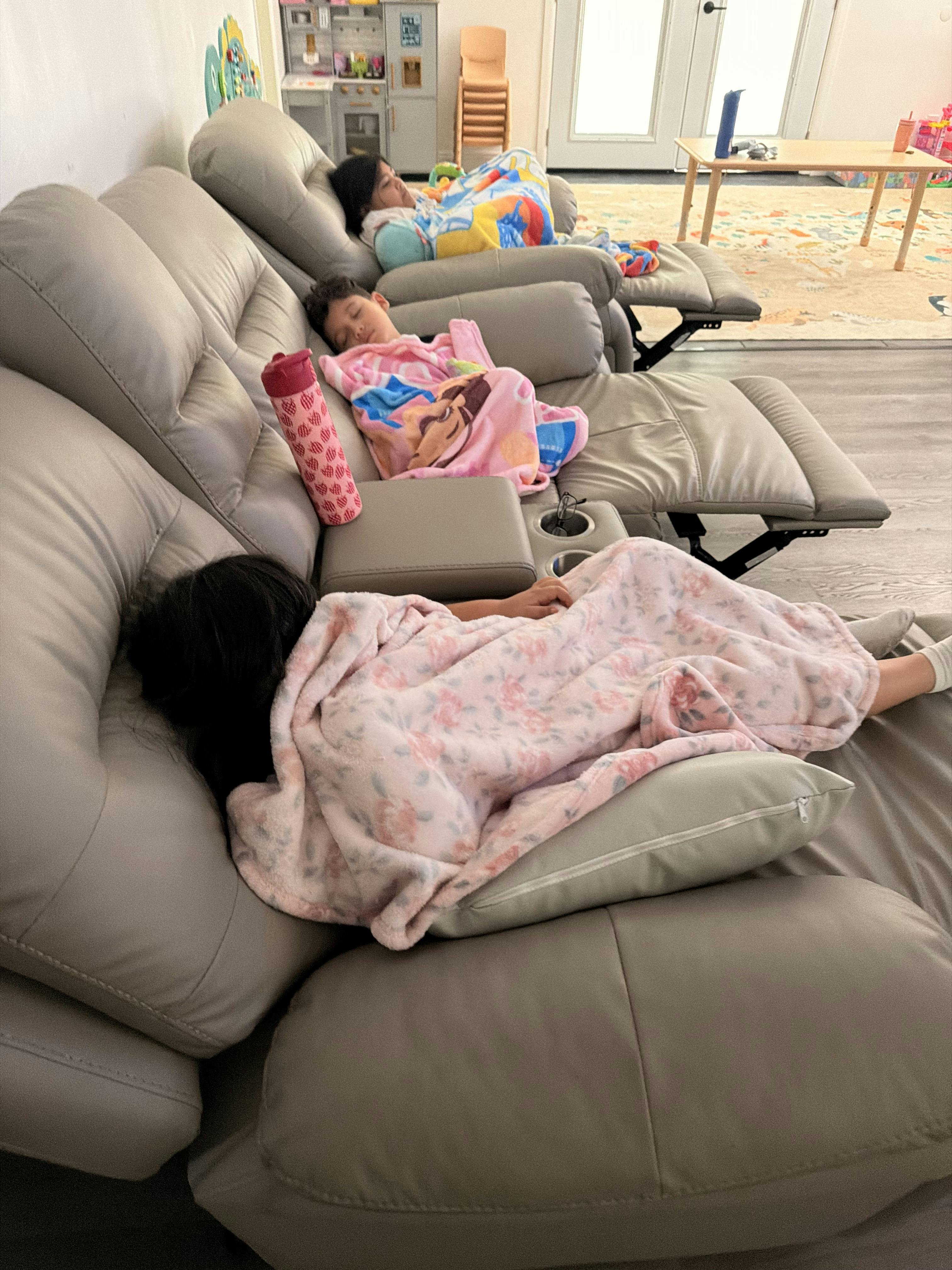 Three children sleeping under blankets on a reclining couch