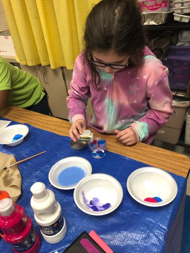 Child pouring liquid into a small cup on a table with paint supplies