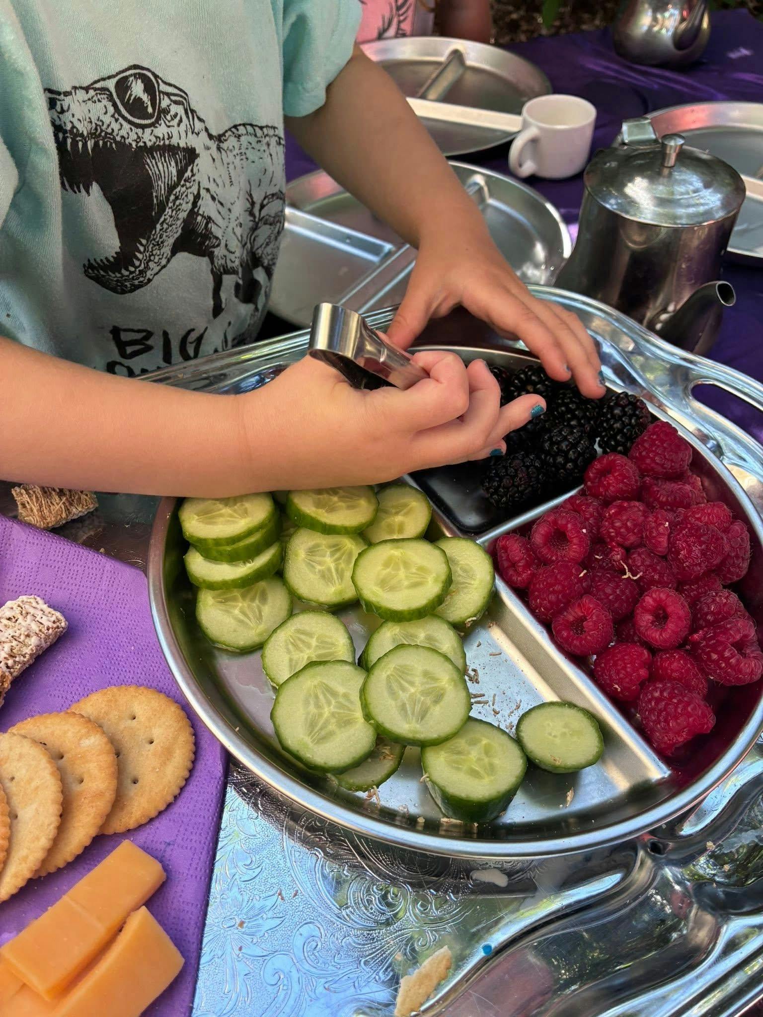 Child using tongs to pick up a blackberry from a tray of fruit and vegetables