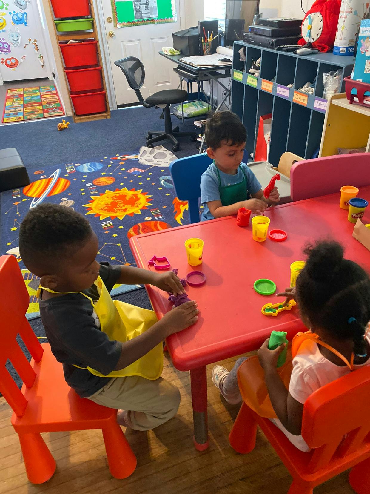 Three children playing with modeling clay at a red table in a classroom