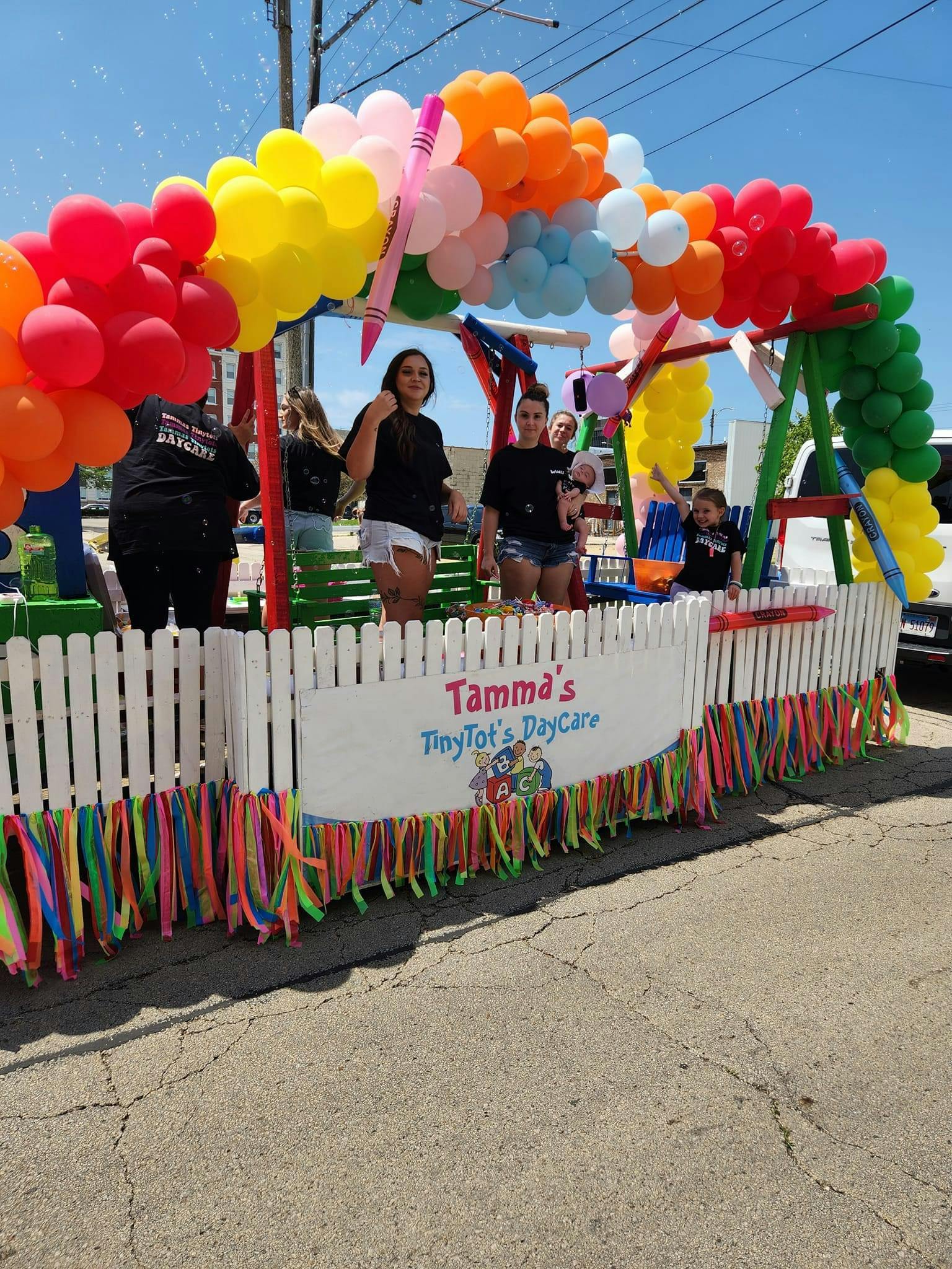 People on a parade float decorated with balloons and a sign for "Tamma's TinyTot's Daycare"
