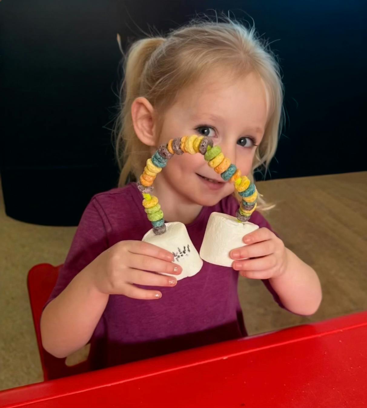 Child holding a craft made from cereal on string and marshmallow cups