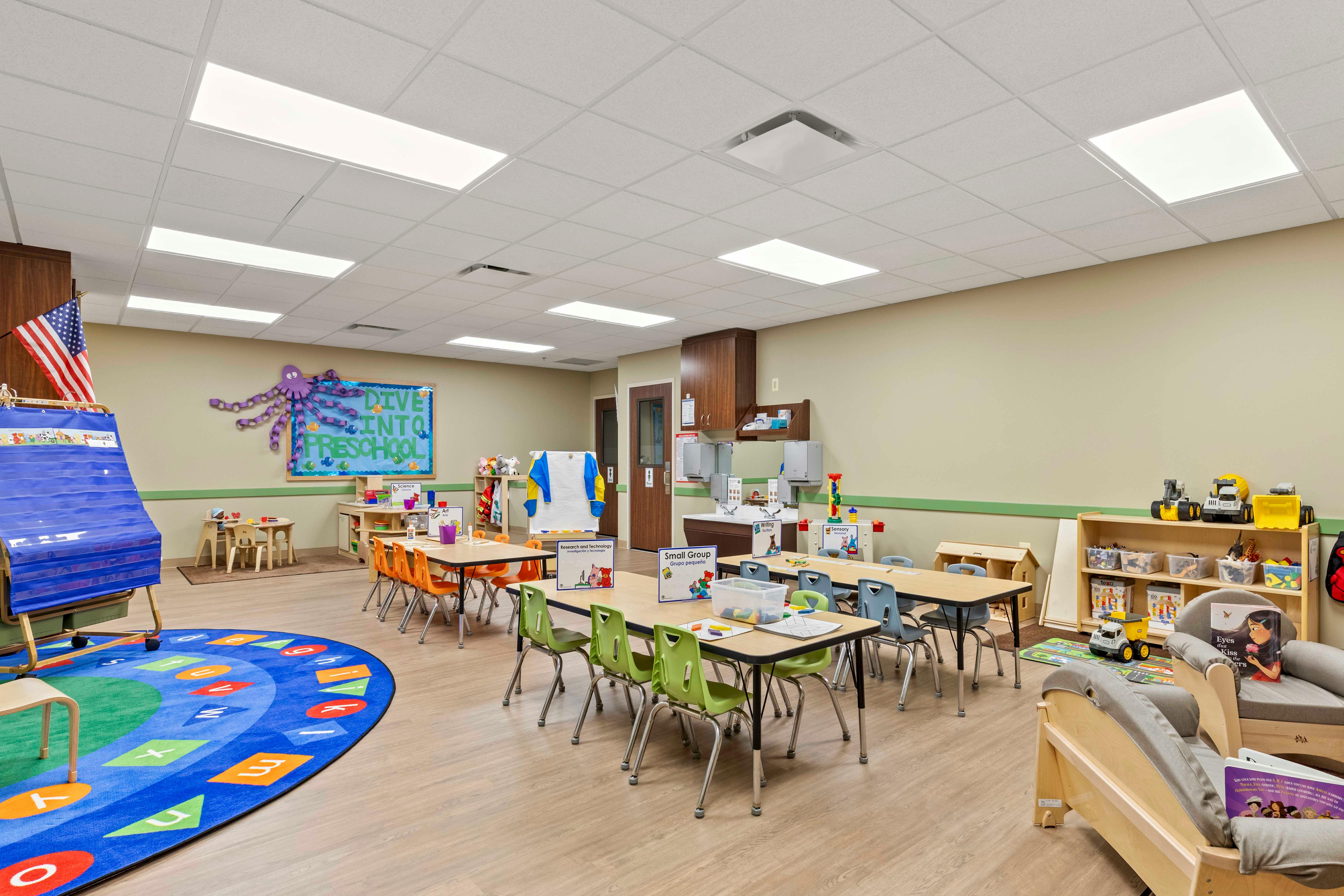 An empty preschool classroom with tables, chairs, a rug with letters, and learning stations