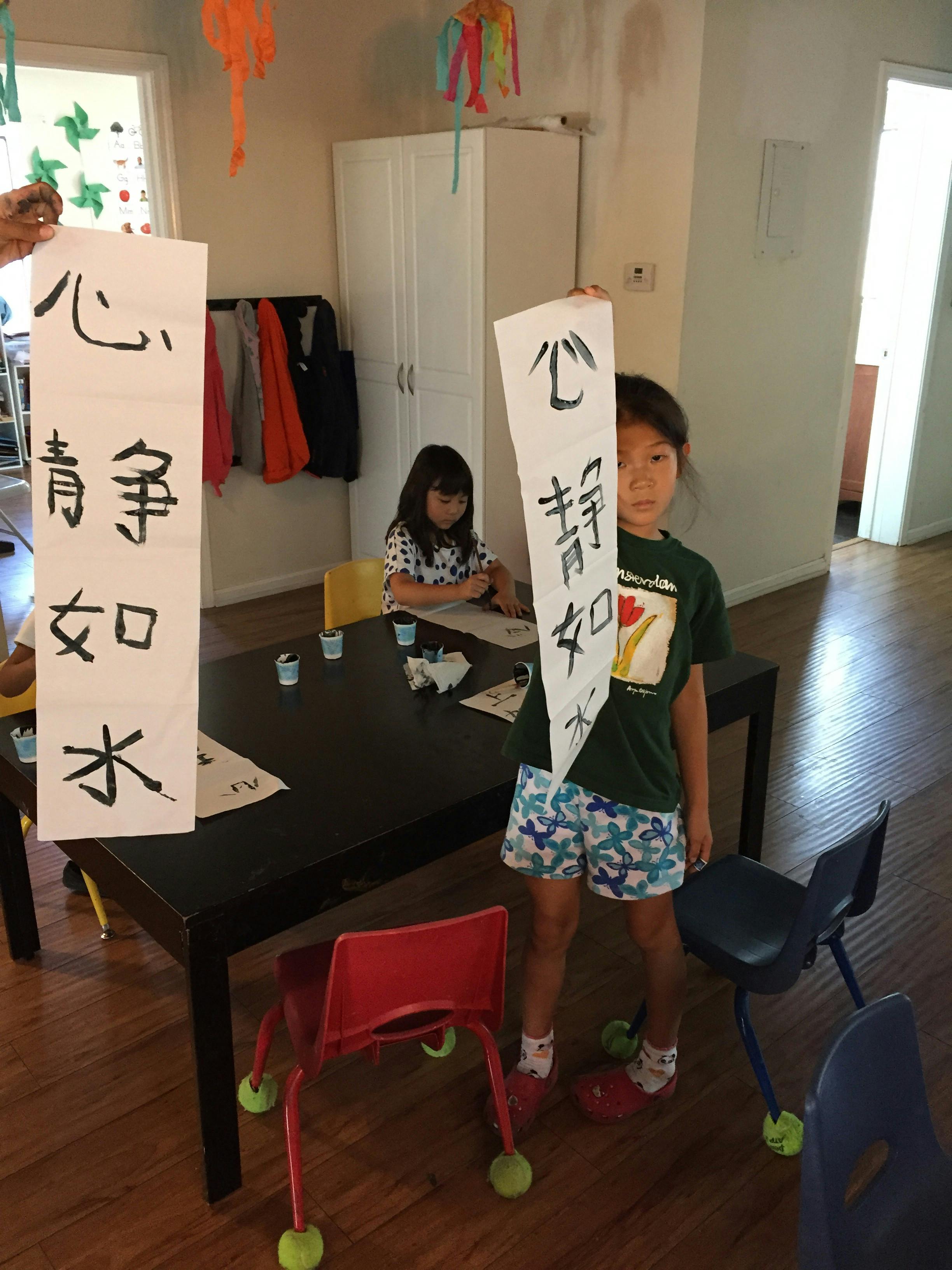 Children doing calligraphy, one holding up a paper with characters
