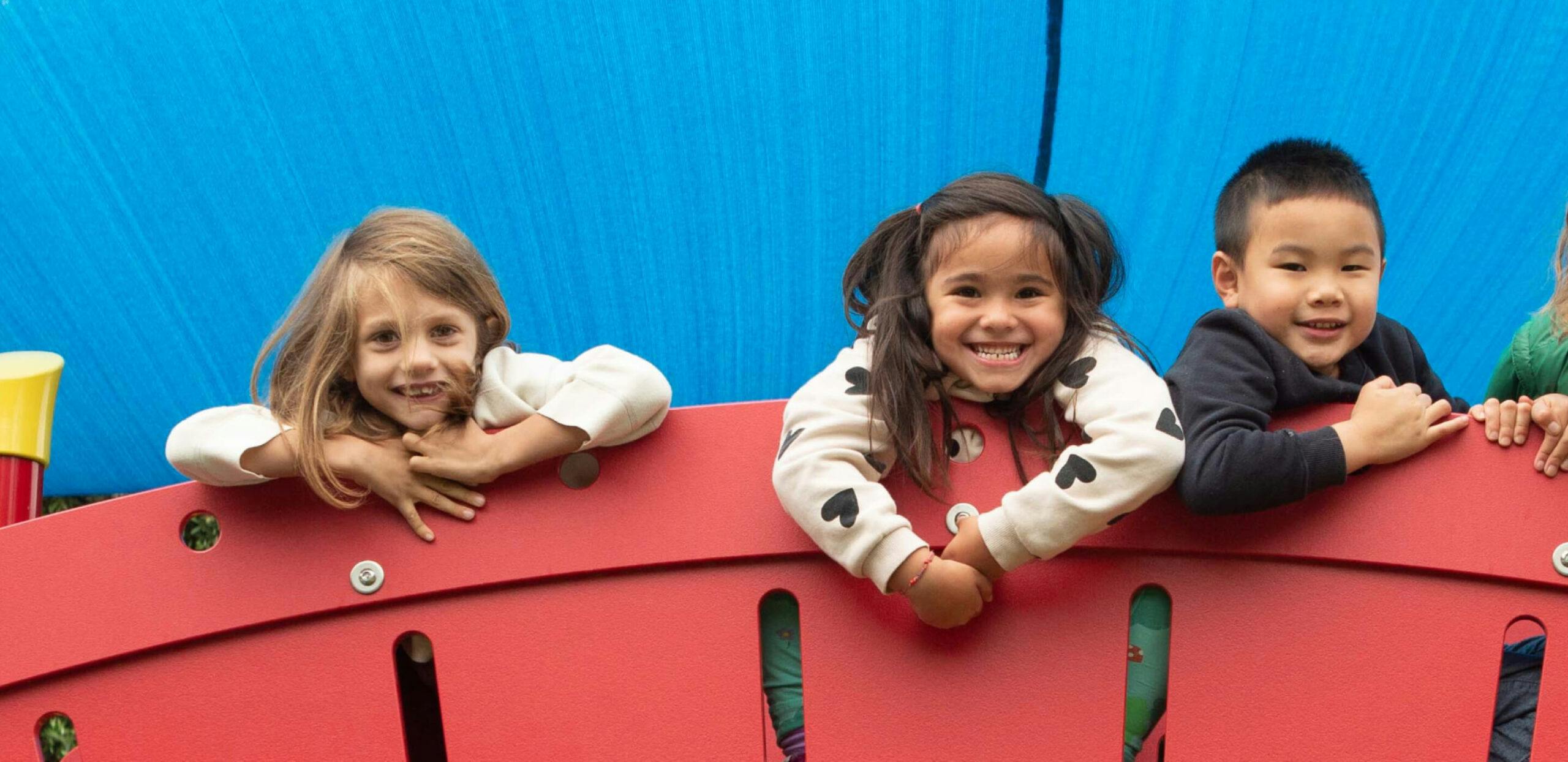 Three children peeking over a red playground structure
