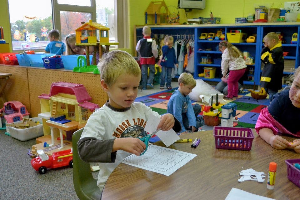 Child cutting paper at a table in a classroom with other children playing