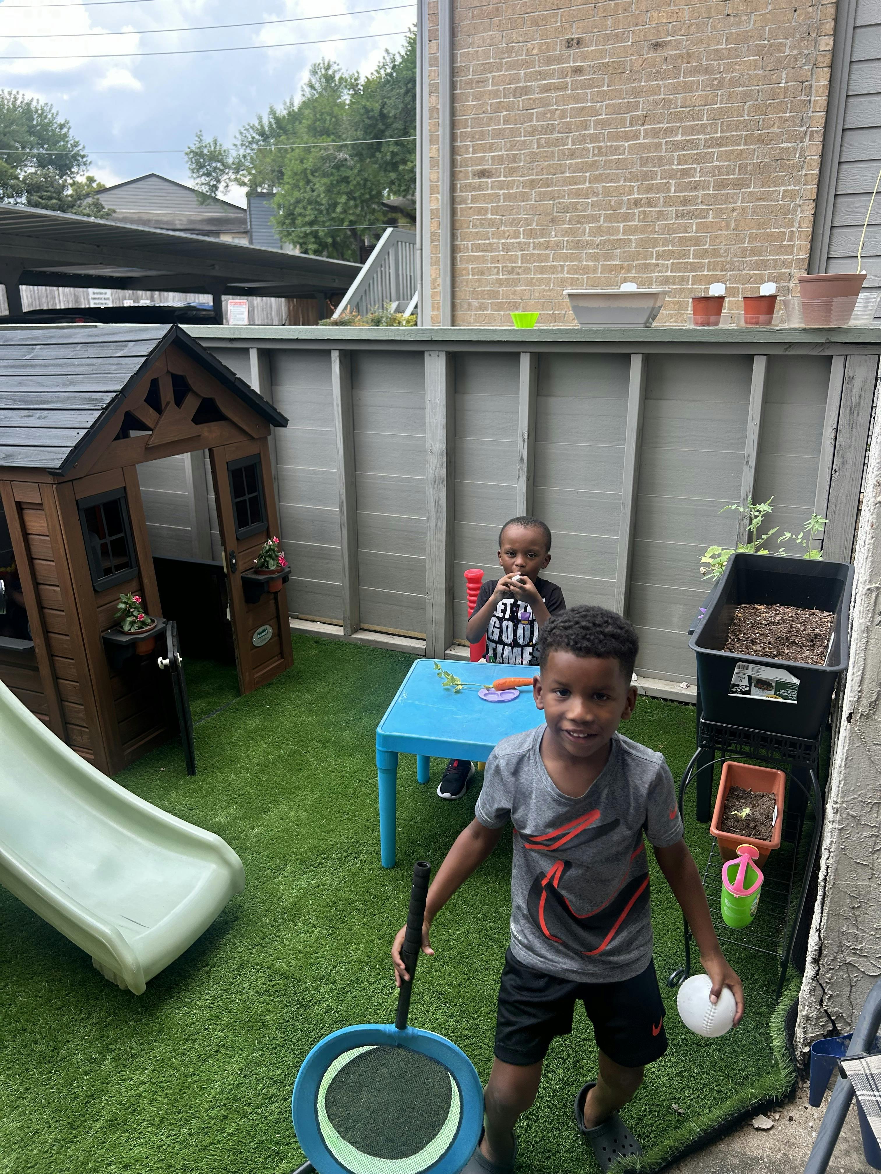 Two children in a backyard, one holding a racket and ball, another sitting at a table