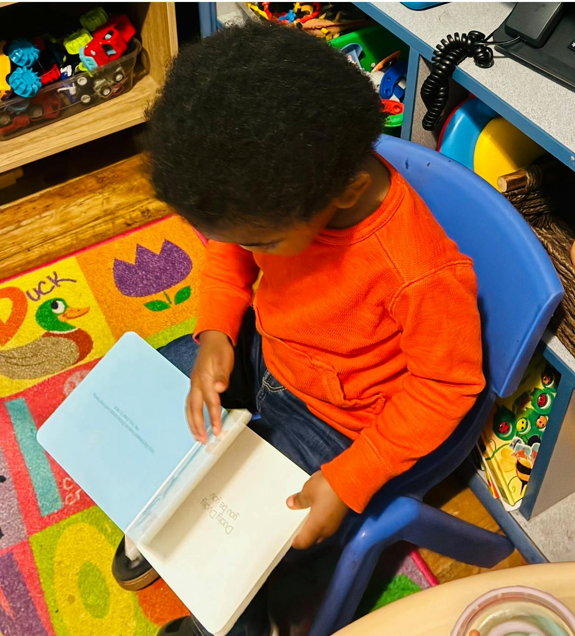 Child in orange shirt sitting on a blue chair and looking at a book