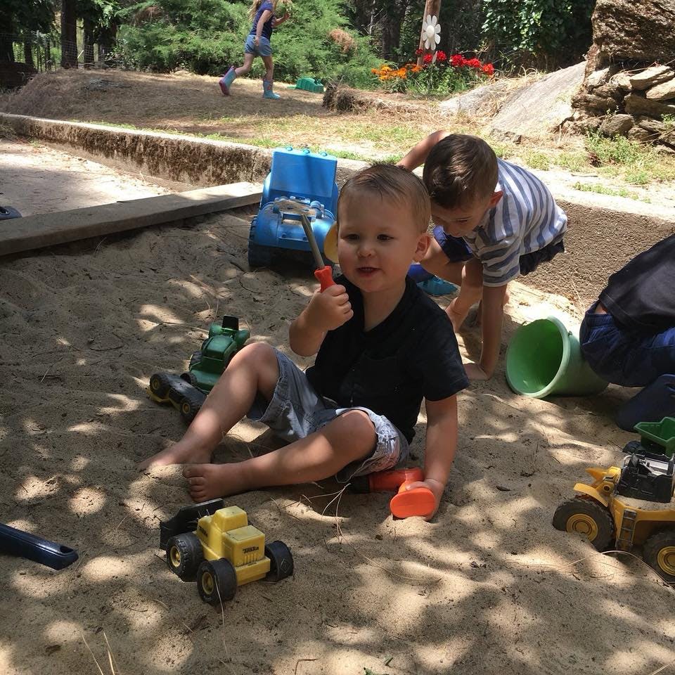 Child sitting in sandbox holding a toy, another child playing in background