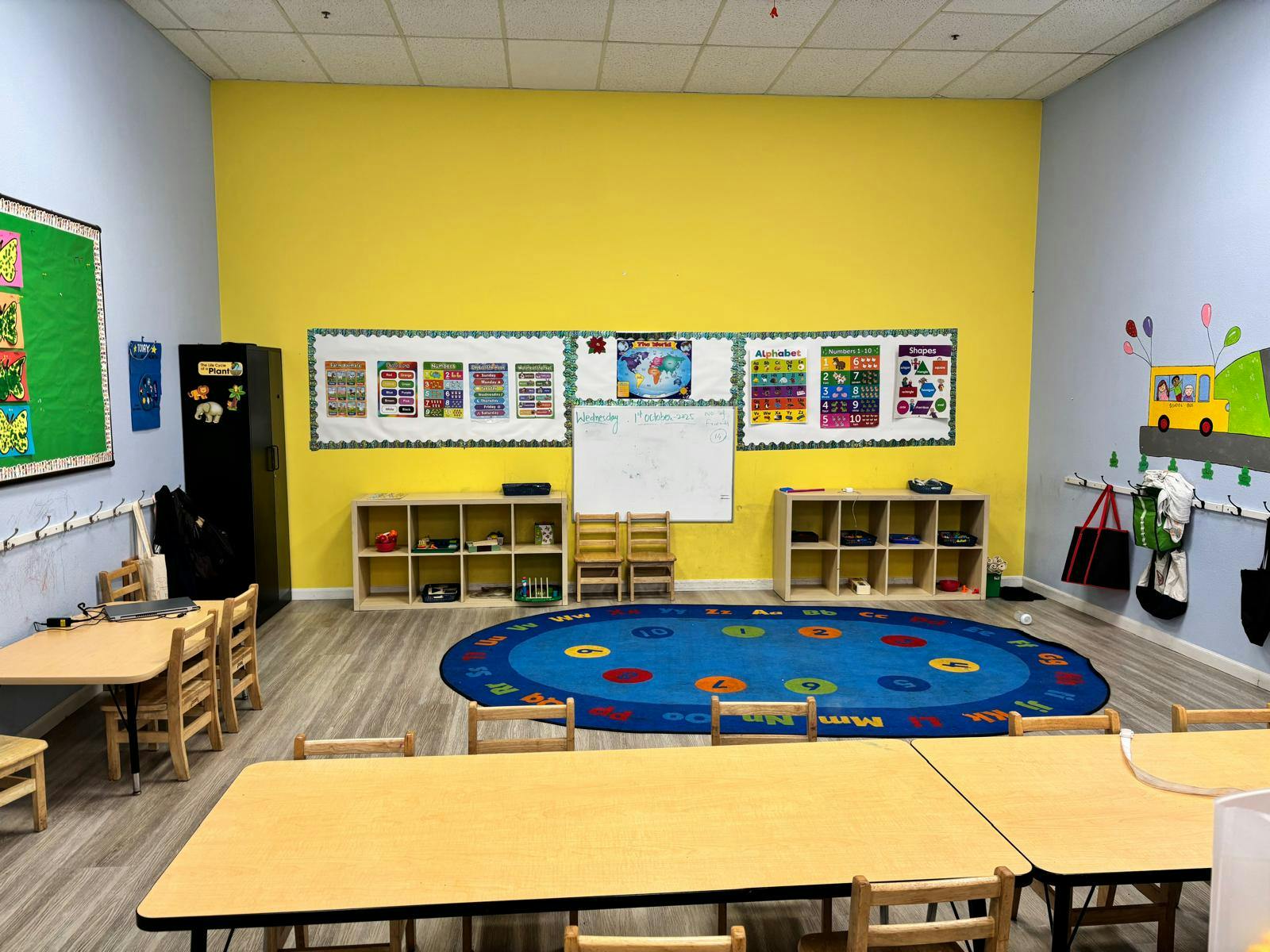 Empty classroom with tables, chairs, colorful rug, and educational posters on a yellow wall