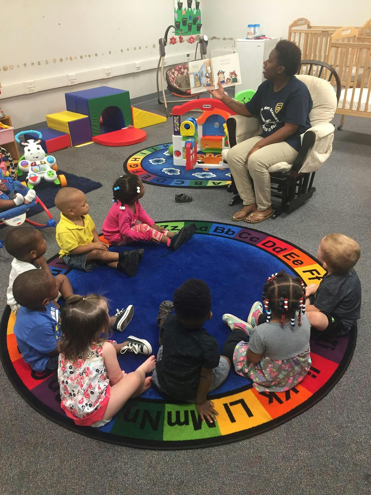 Person reading to a group of children sitting on a colorful alphabet rug