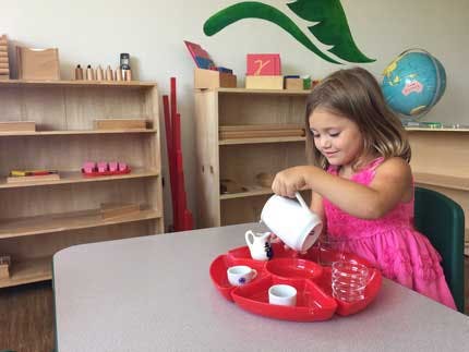 Child pouring water from a pitcher into a cup at a table in a classroom