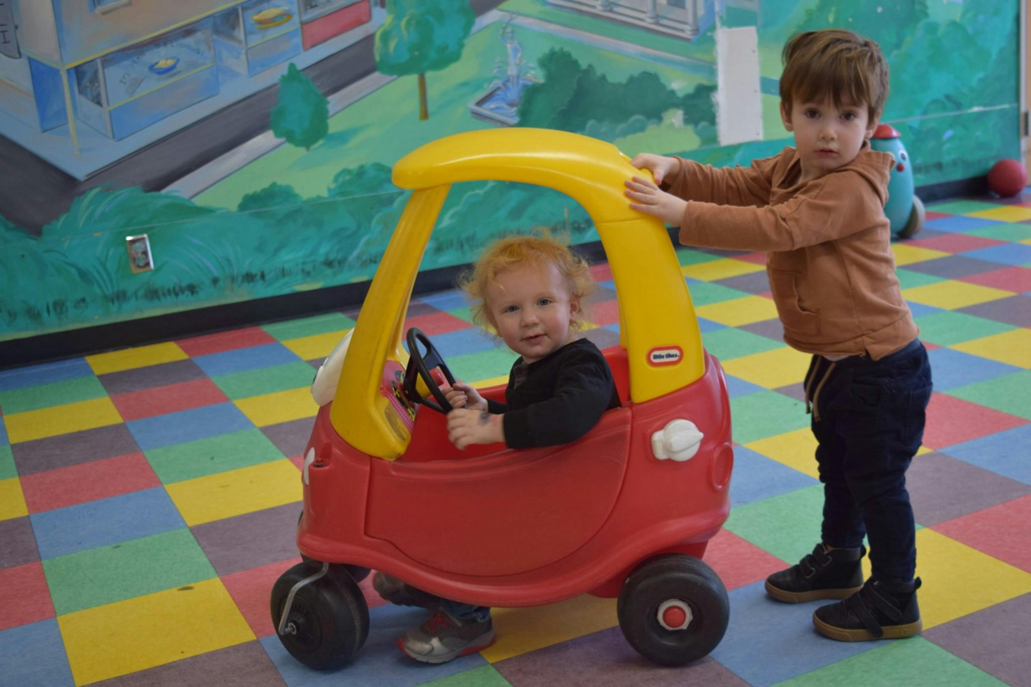 Child in toy car with another child pushing it