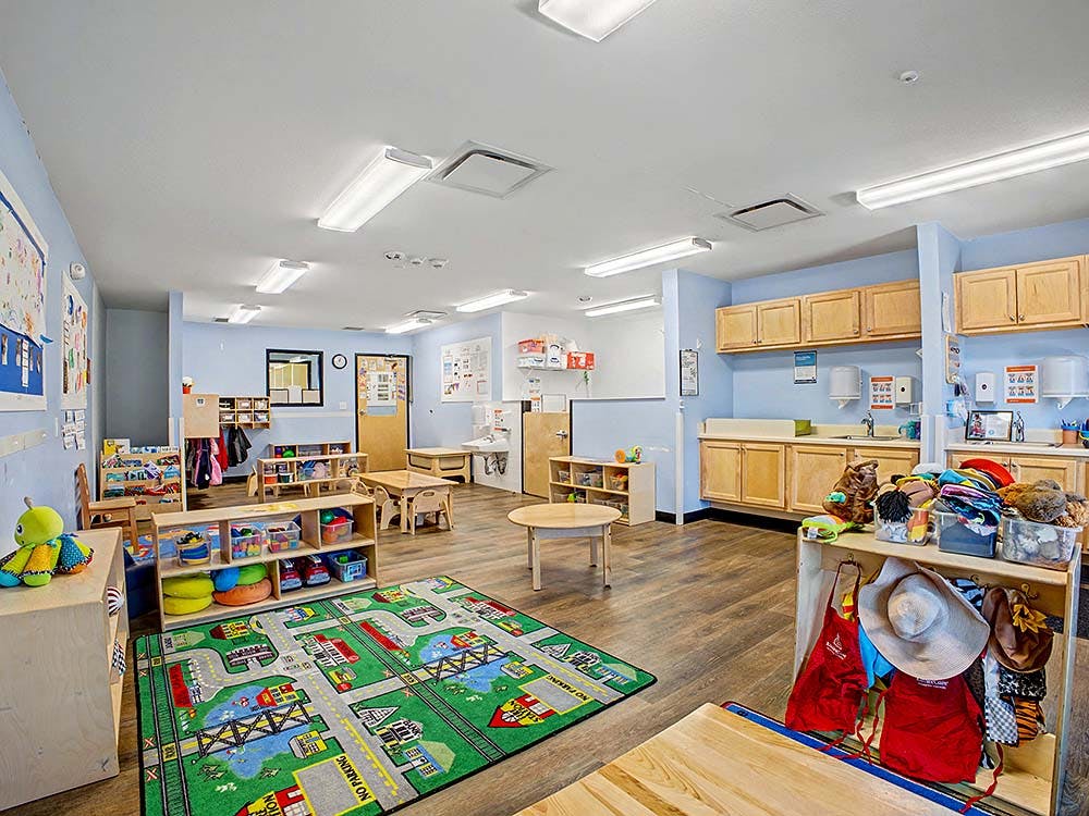 A classroom with small tables and chairs, a play rug, and storage shelves