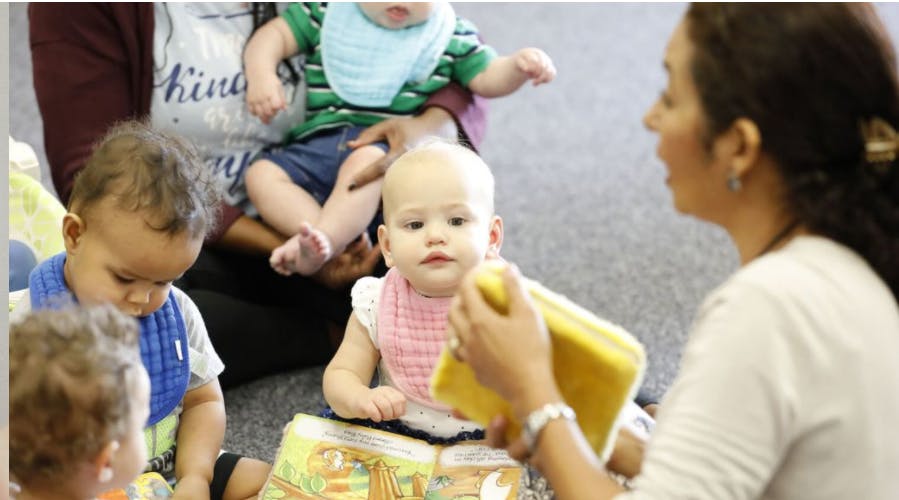 Adult holding a yellow object with multiple babies and an adult in the background