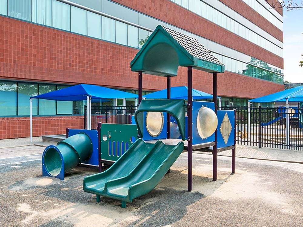 A playground with a green slide and tunnel, blue shade covers, and a brick building