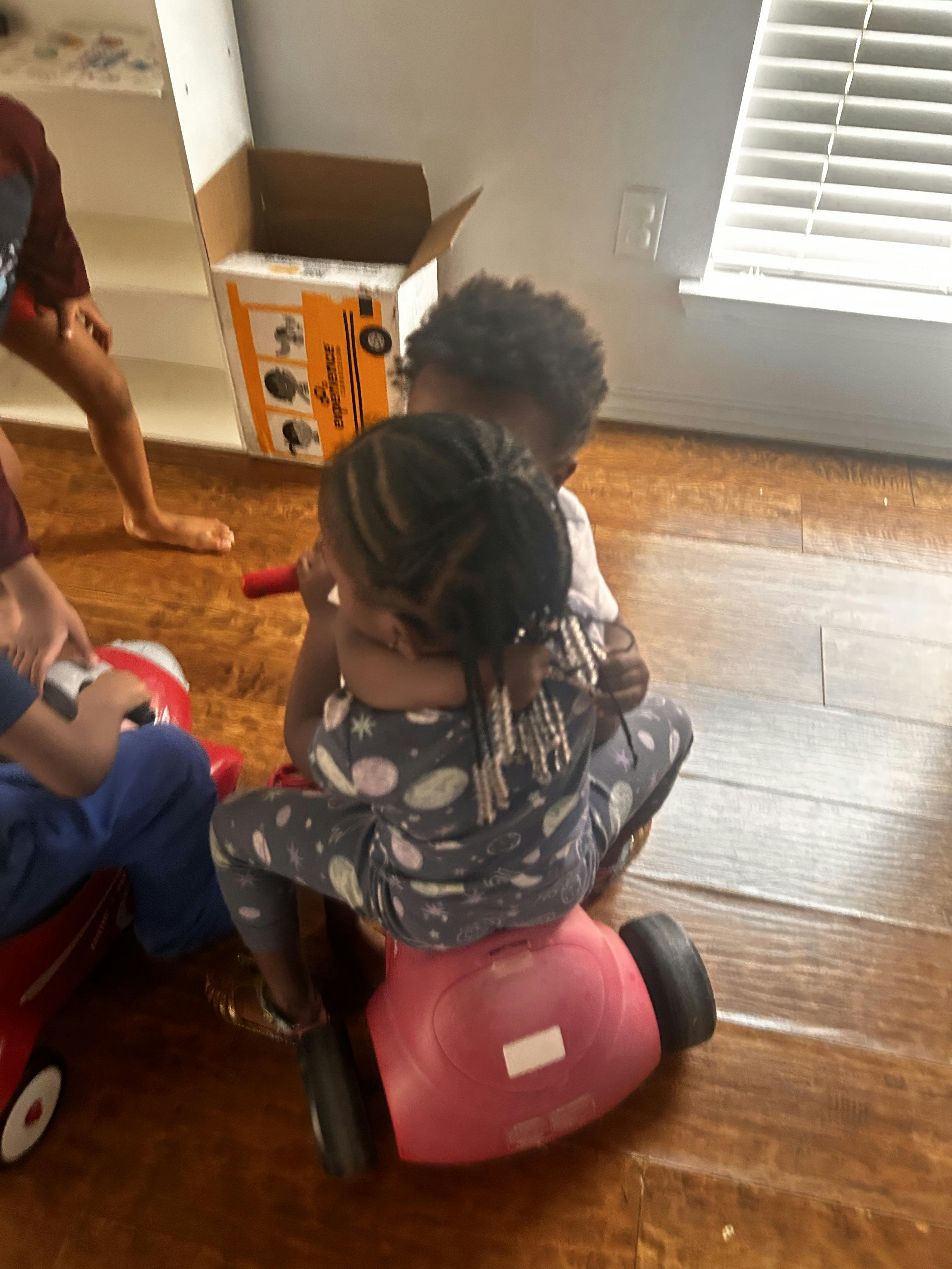 Two children hugging while sitting on a toy ride-on vehicle indoors