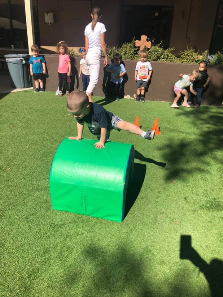 Child climbing over a green cylinder on artificial grass with other children nearby