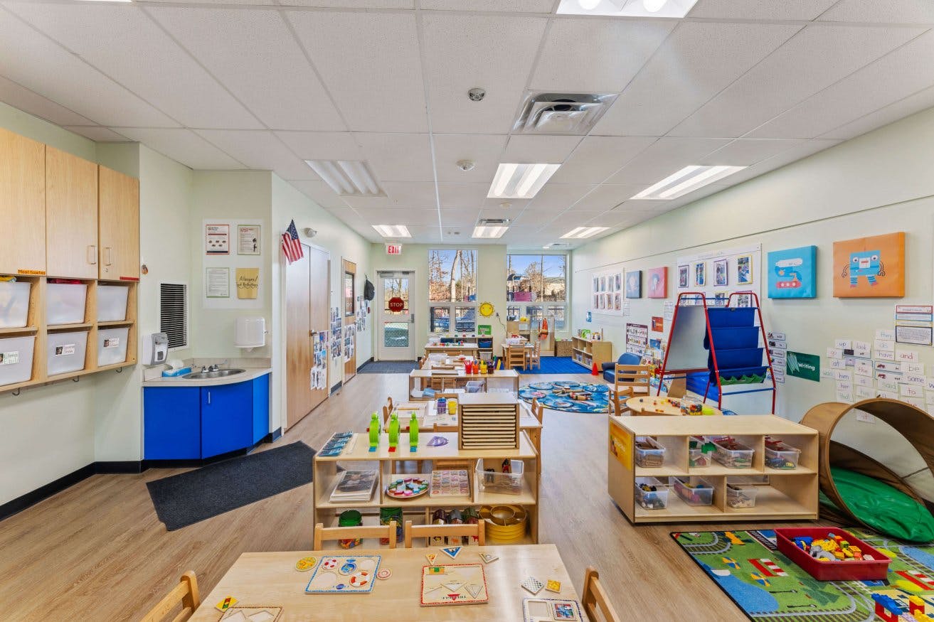 Classroom with child-sized tables and chairs, shelves with toys, and a sink area