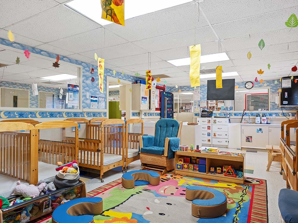 Childcare room with multiple cribs, a blue rocking chair, and colorful play rug