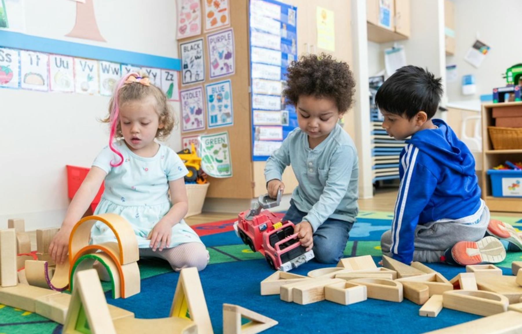 Three children playing on a rug with wooden blocks and a toy truck