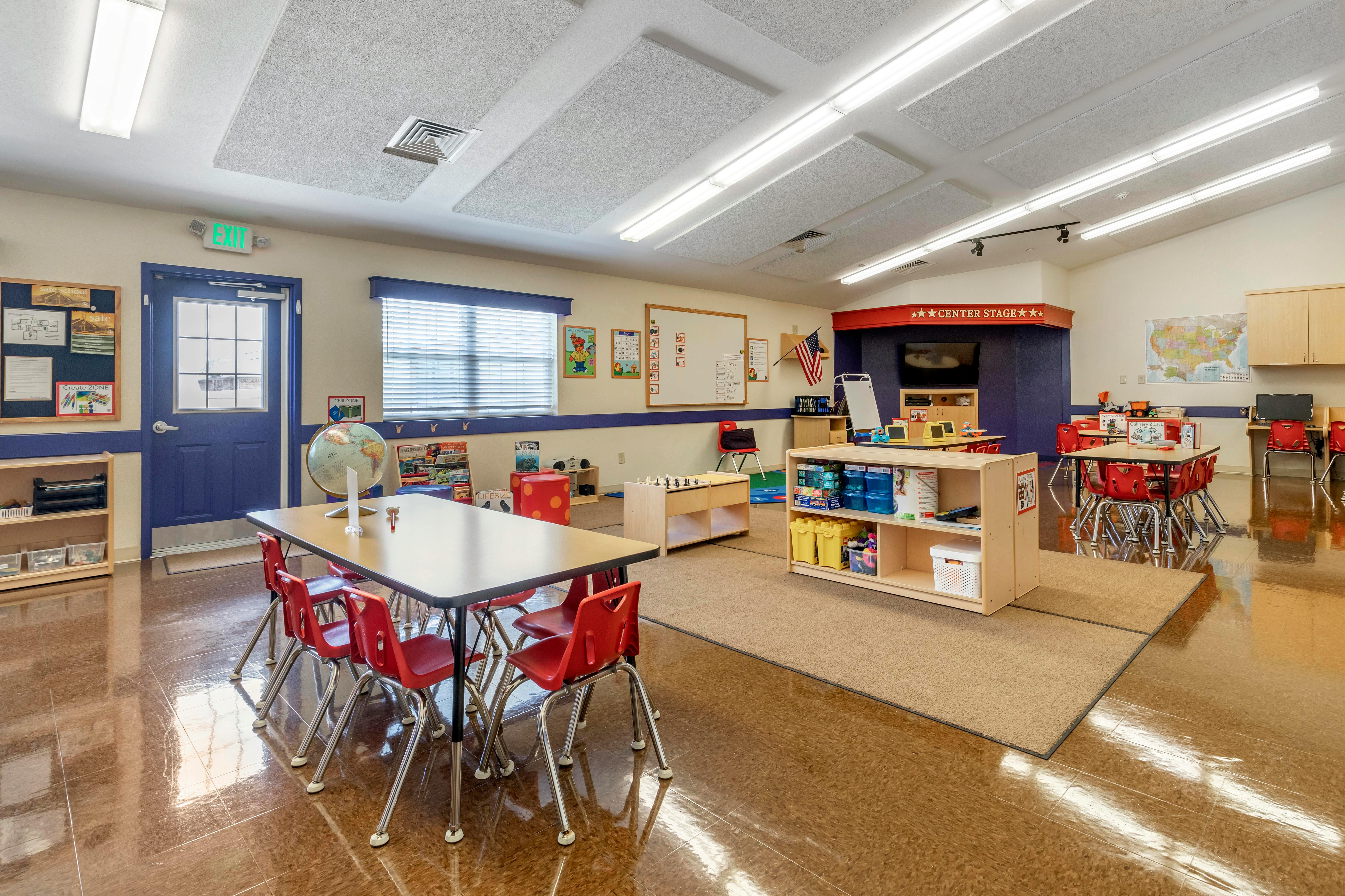 Classroom with small tables and red chairs, play area with rug and toys