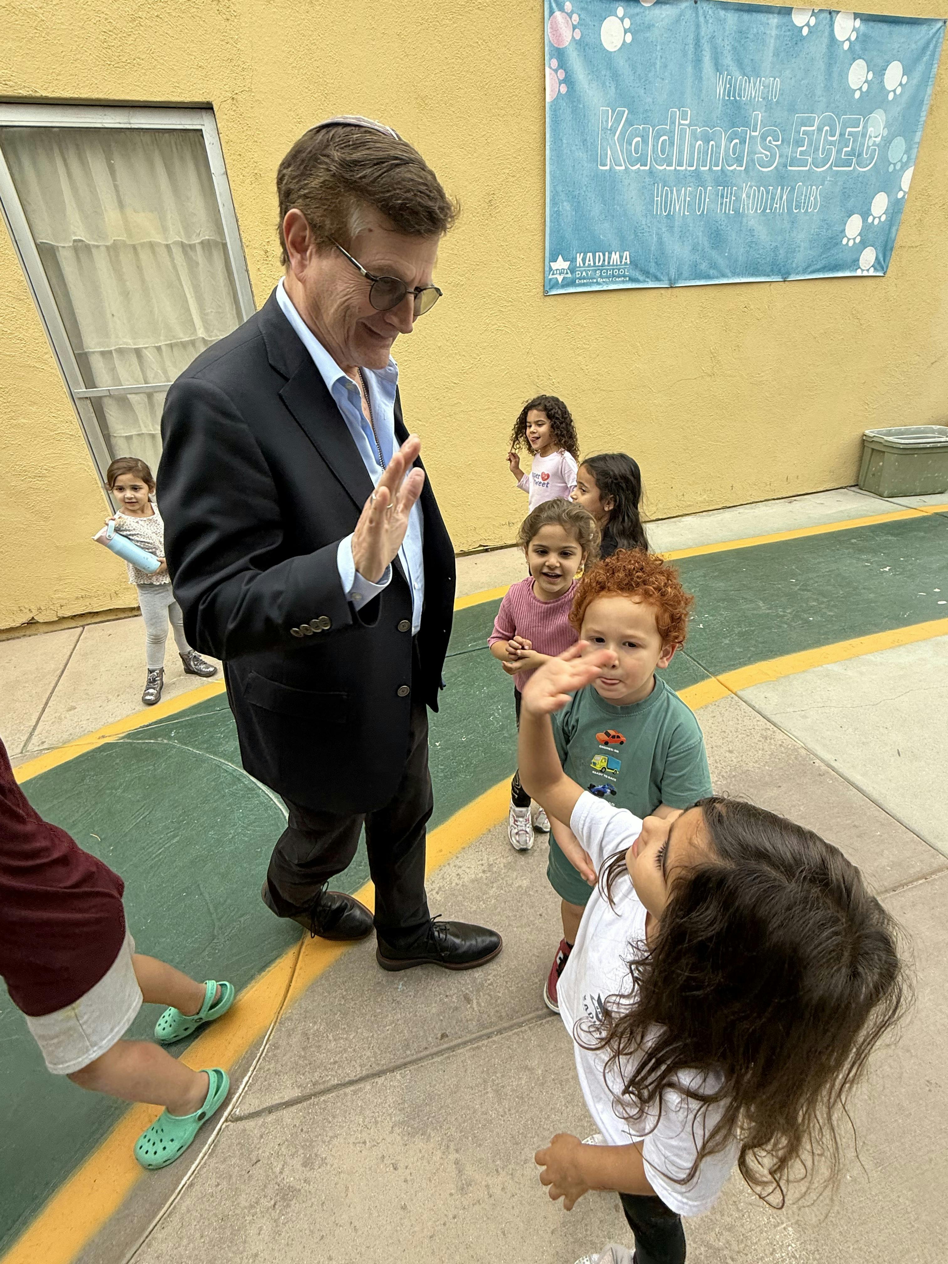 A man in a suit with children, banner says "Welcome to Kadima's ECEC"