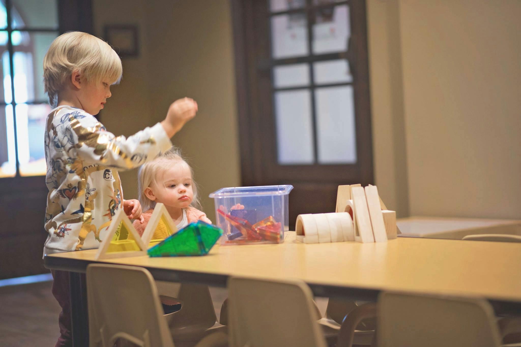 Child standing next to a table with another child sitting at the table