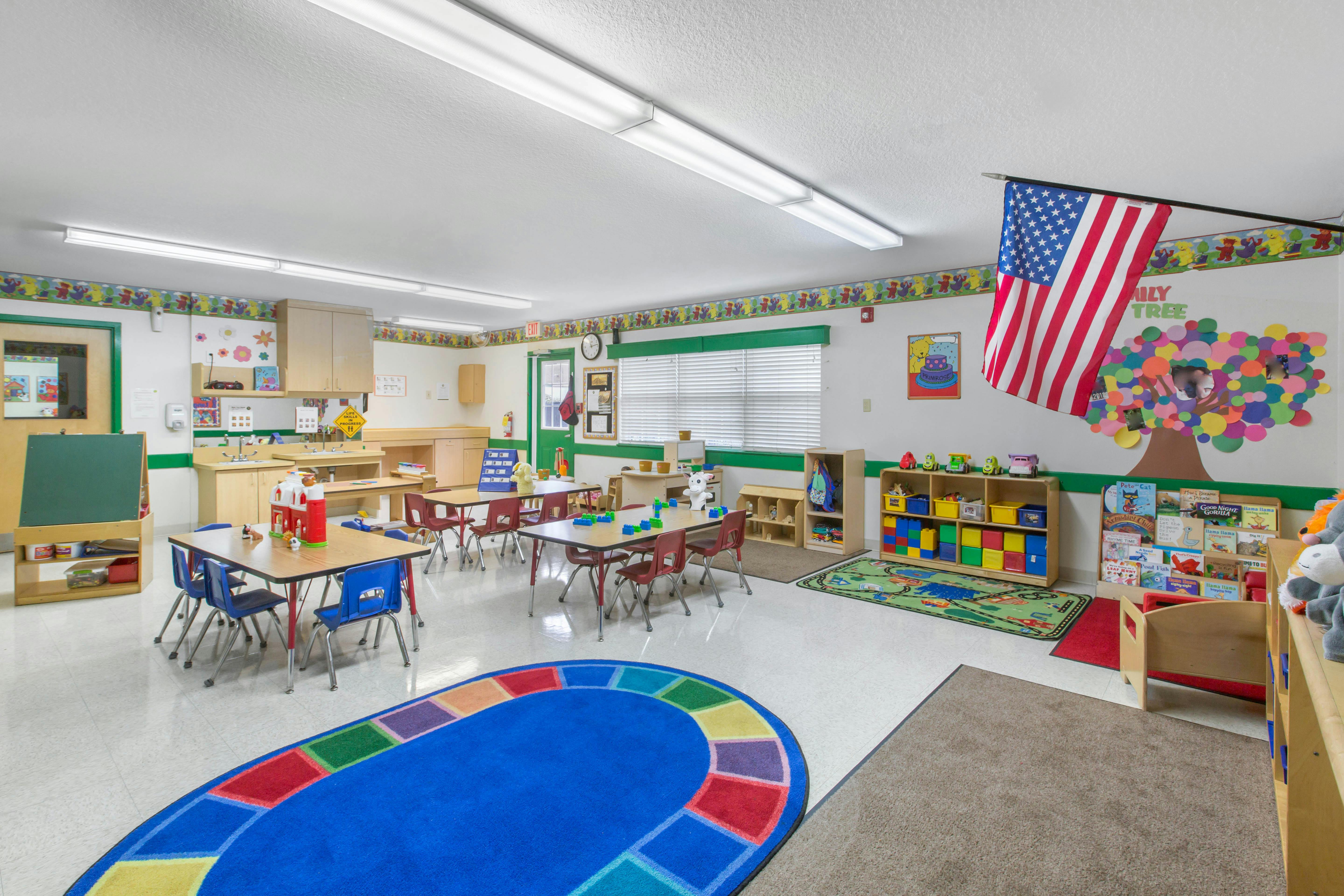 An empty childcare classroom with small tables and chairs, toys, and American flag