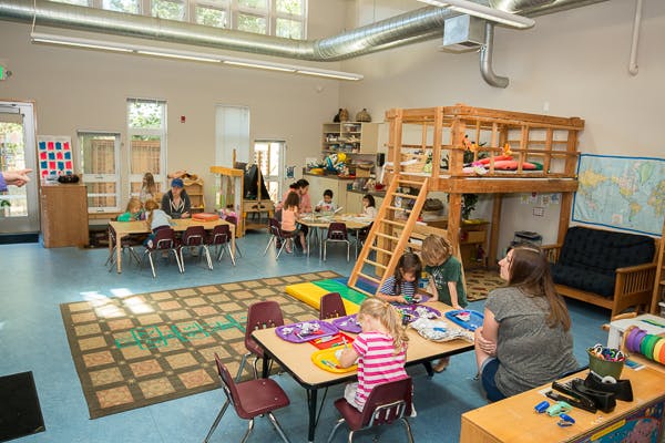 People and children in a classroom with tables and play structures