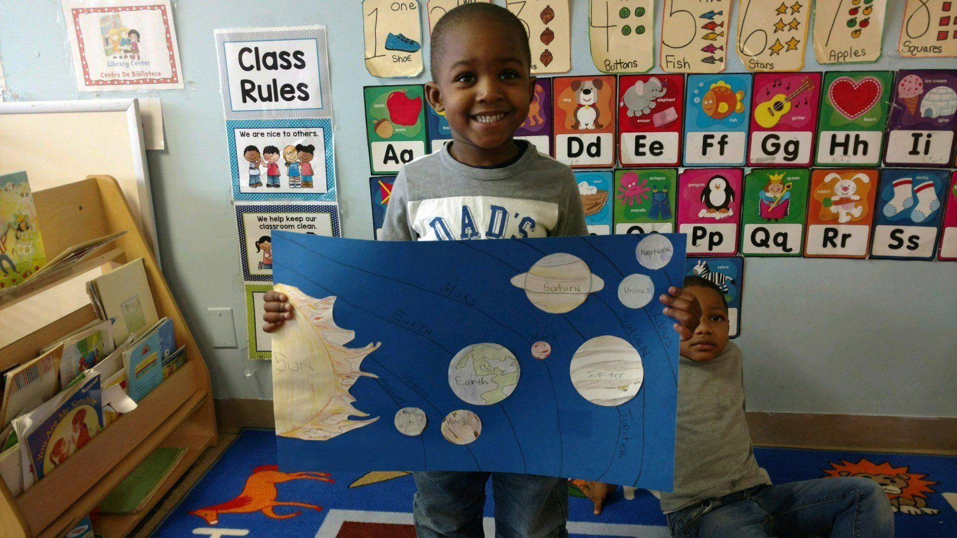 Child in a classroom holding up a solar system poster, smiling at the camera