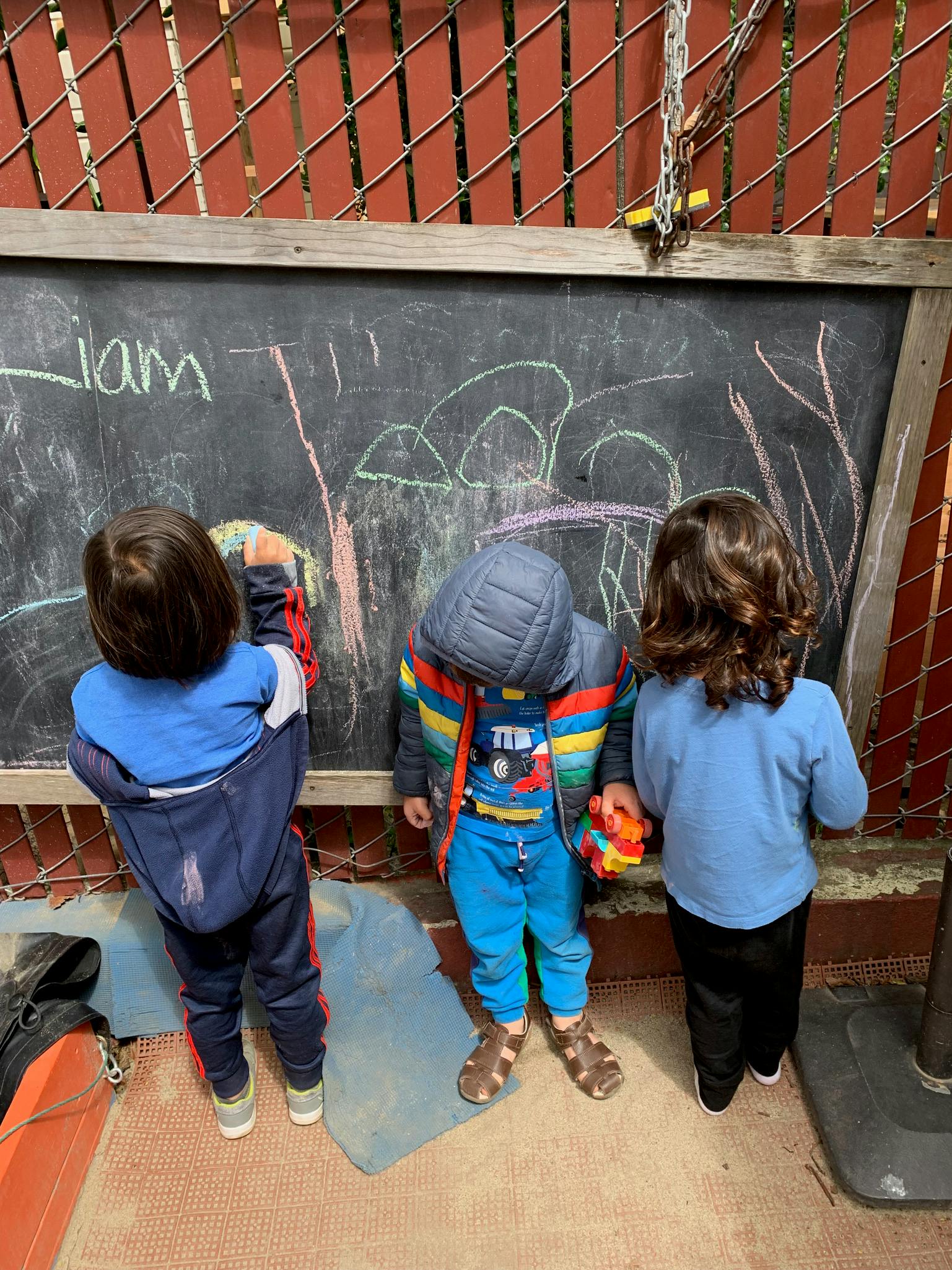 Three children drawing on a large outdoor chalkboard