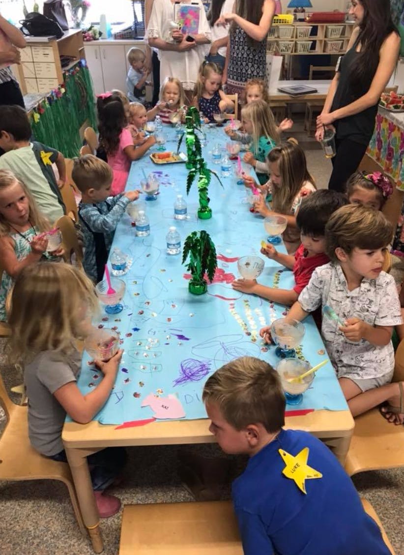 Children sitting at a long table with adults standing adults nearby