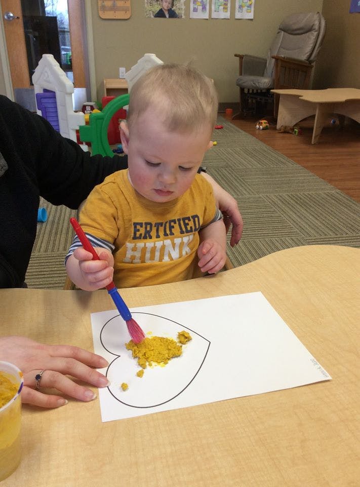 Child using a brush to spread yellow material on a heart outline