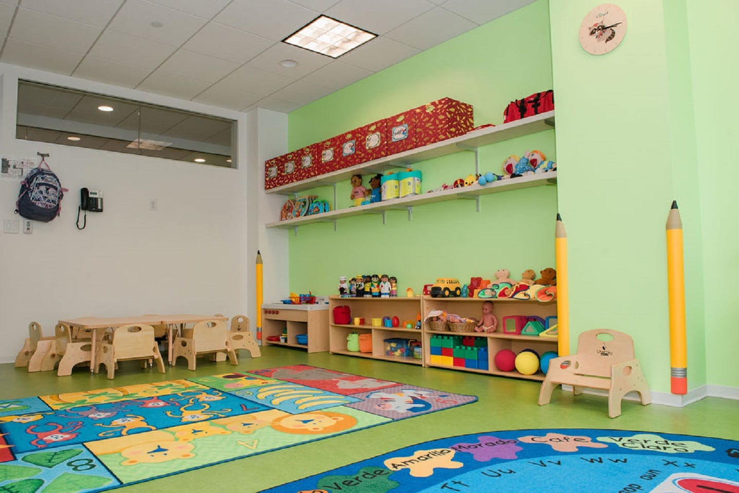 Classroom with small wooden tables and chairs, toys on shelves, and colorful play mats