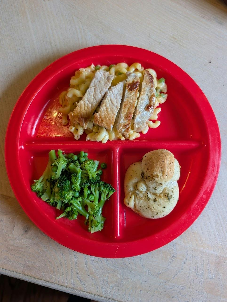 A divided red plate containing sliced meat, macaroni, broccoli, peas, and a roll