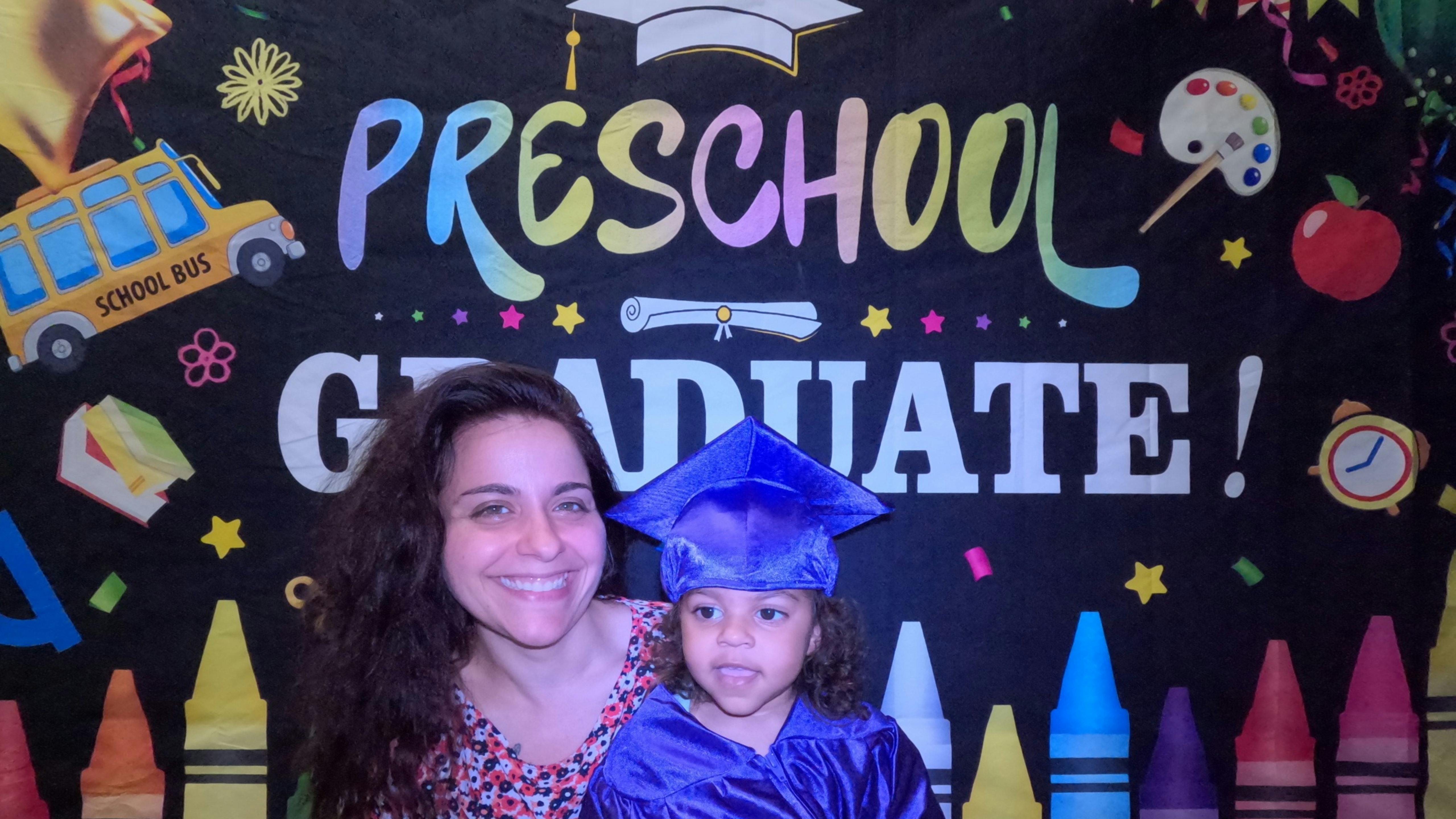 Person and child in graduation cap and gown in front of "Preschool Graduate" sign