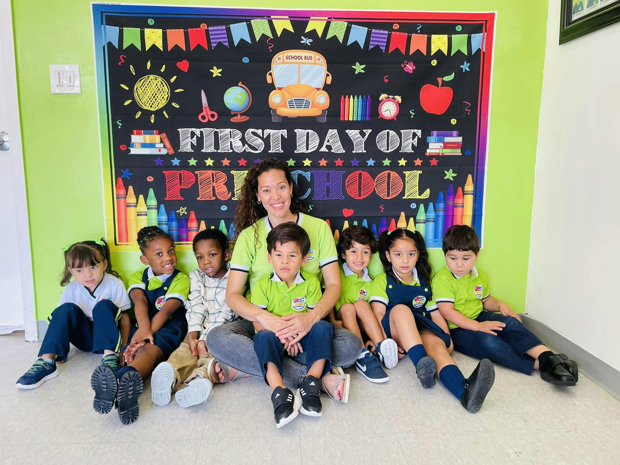 Adult and children sitting in front of a "First Day of Preschool" sign