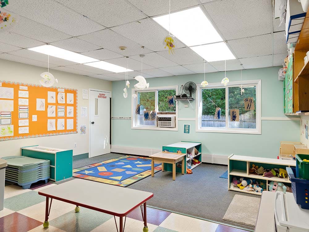 empty classroom with tables, chairs, colorful rug, and windows showing trees