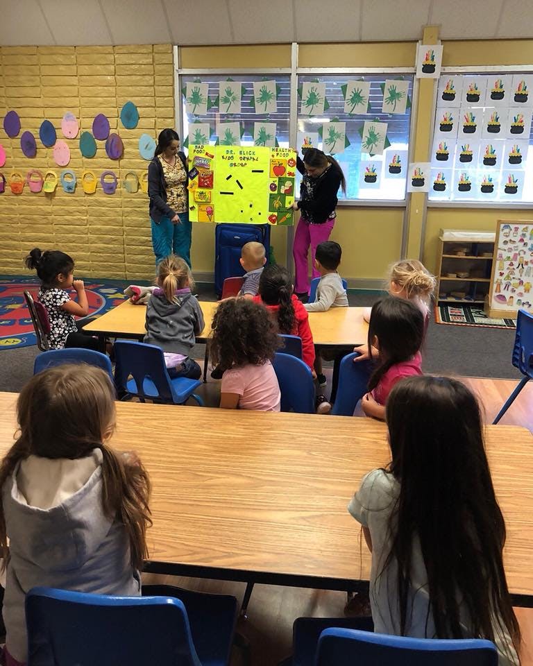 Two people presenting a large yellow poster to a group of children in a classroom