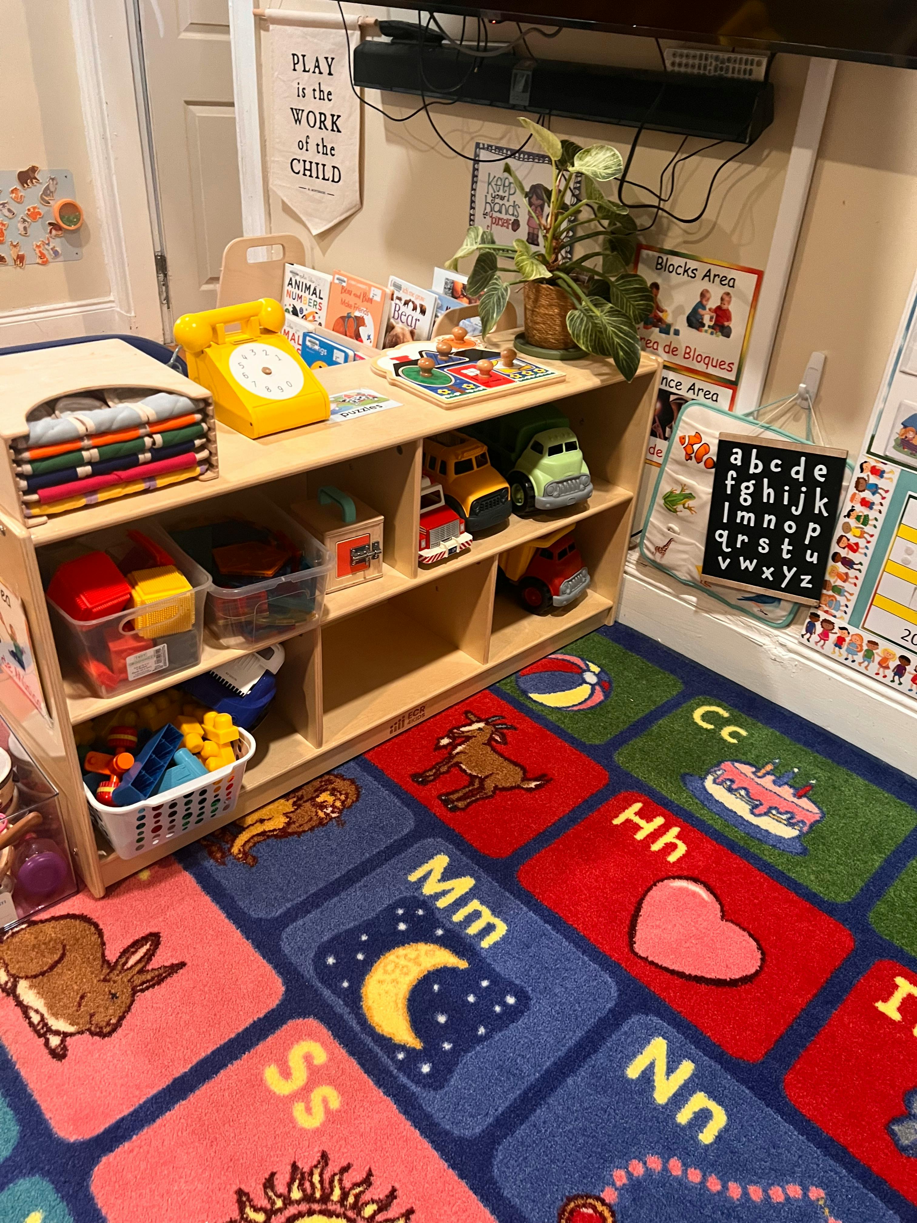 Classroom with a wooden shelf holding toys and books, and an alphabet rug