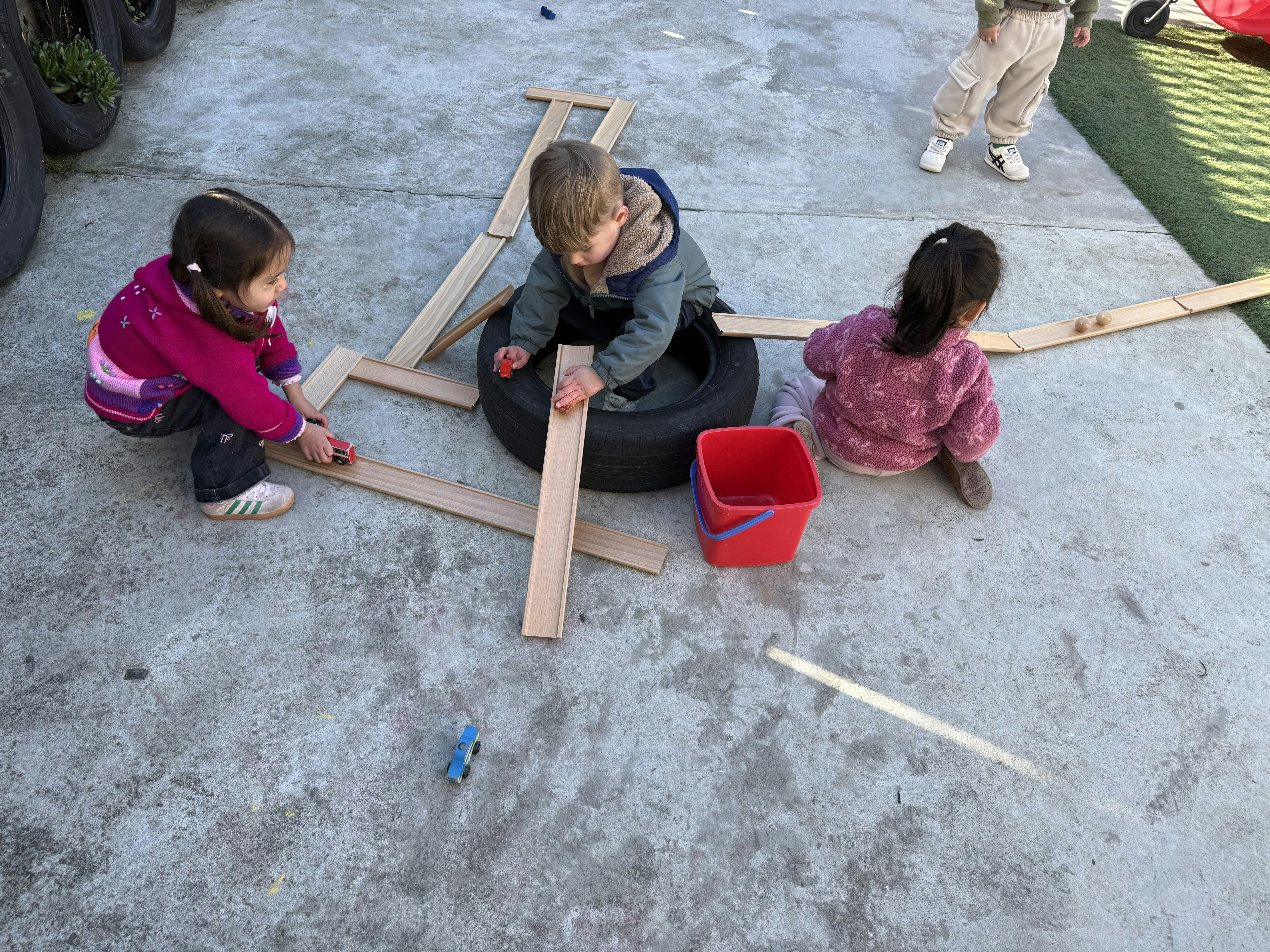 Three children playing with wooden tracks and toy cars on a concrete surface