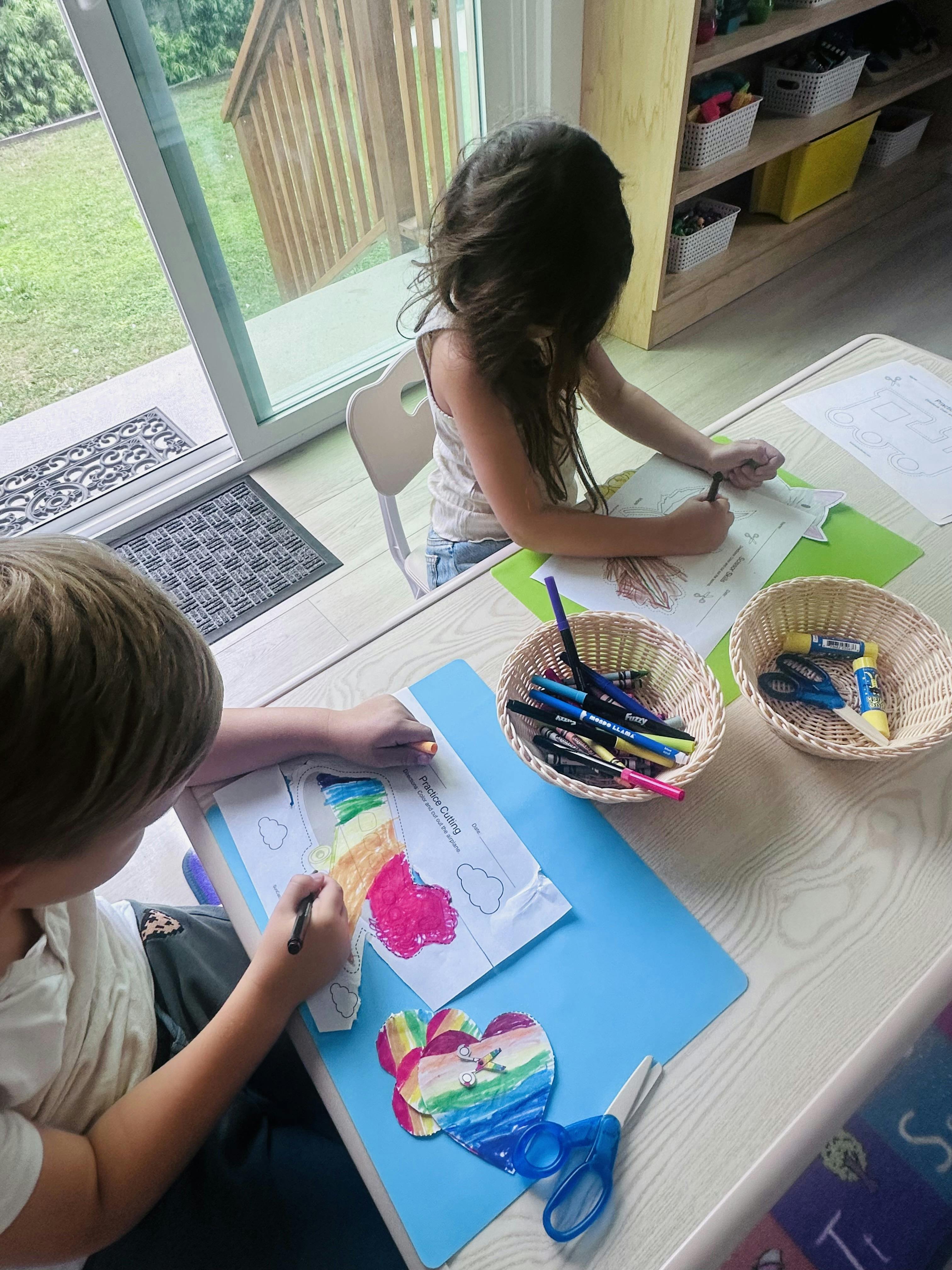 Two children coloring and working on practice cutting worksheets at a table