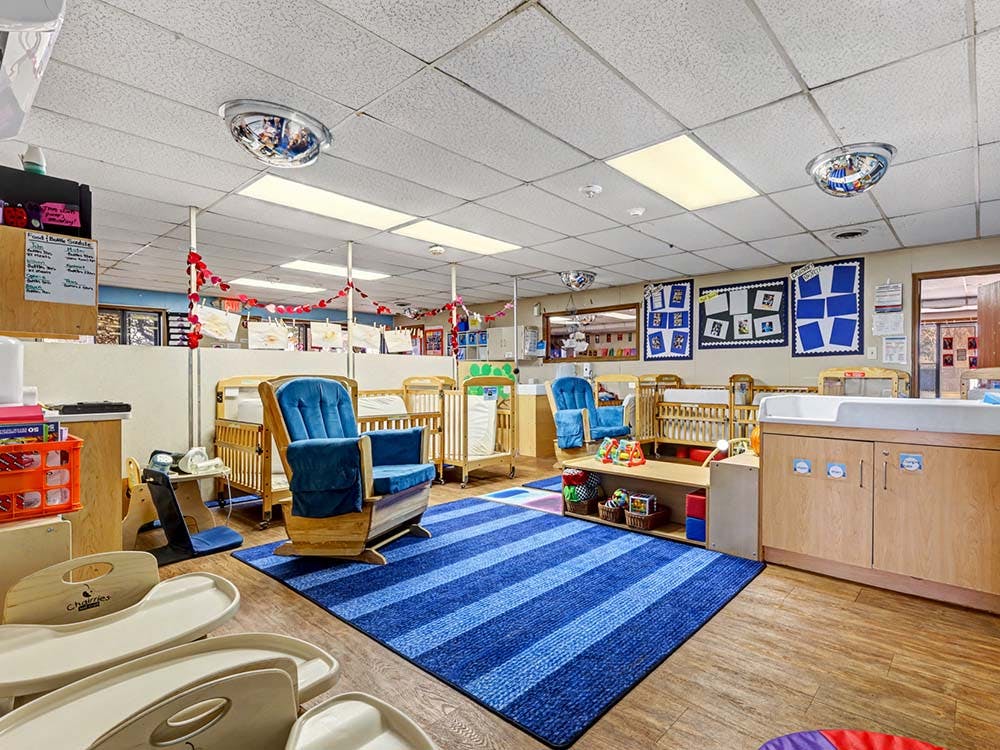 A daycare room with cribs, a blue rocking chair, a changing table, and a blue rug