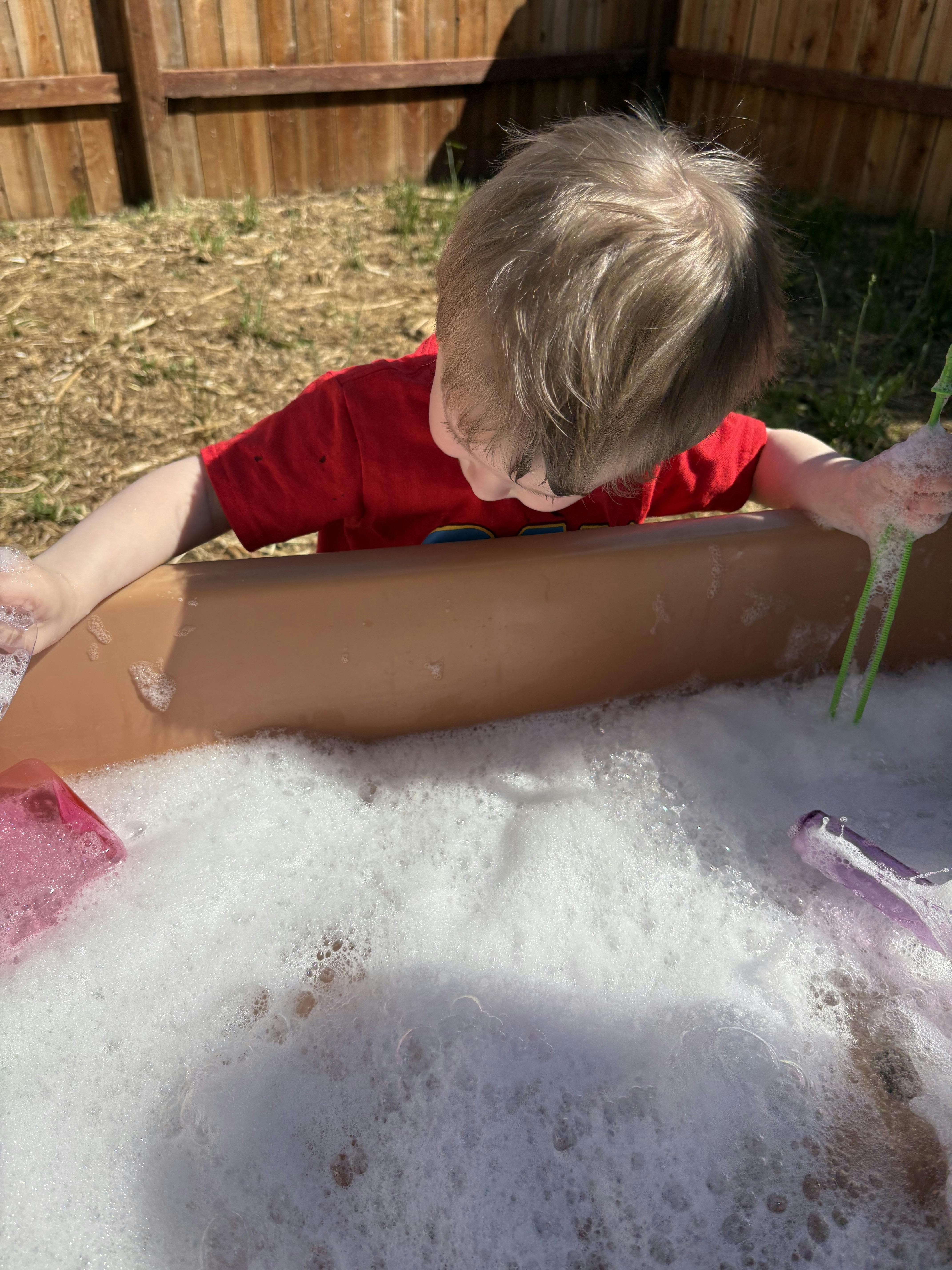 Child in a red shirt playing with bubbles in a brown tub outdoors