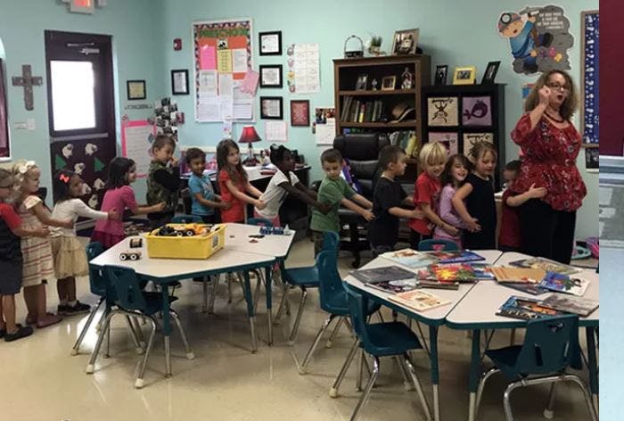 Children holding hands in a line in a classroom with a person standing nearby