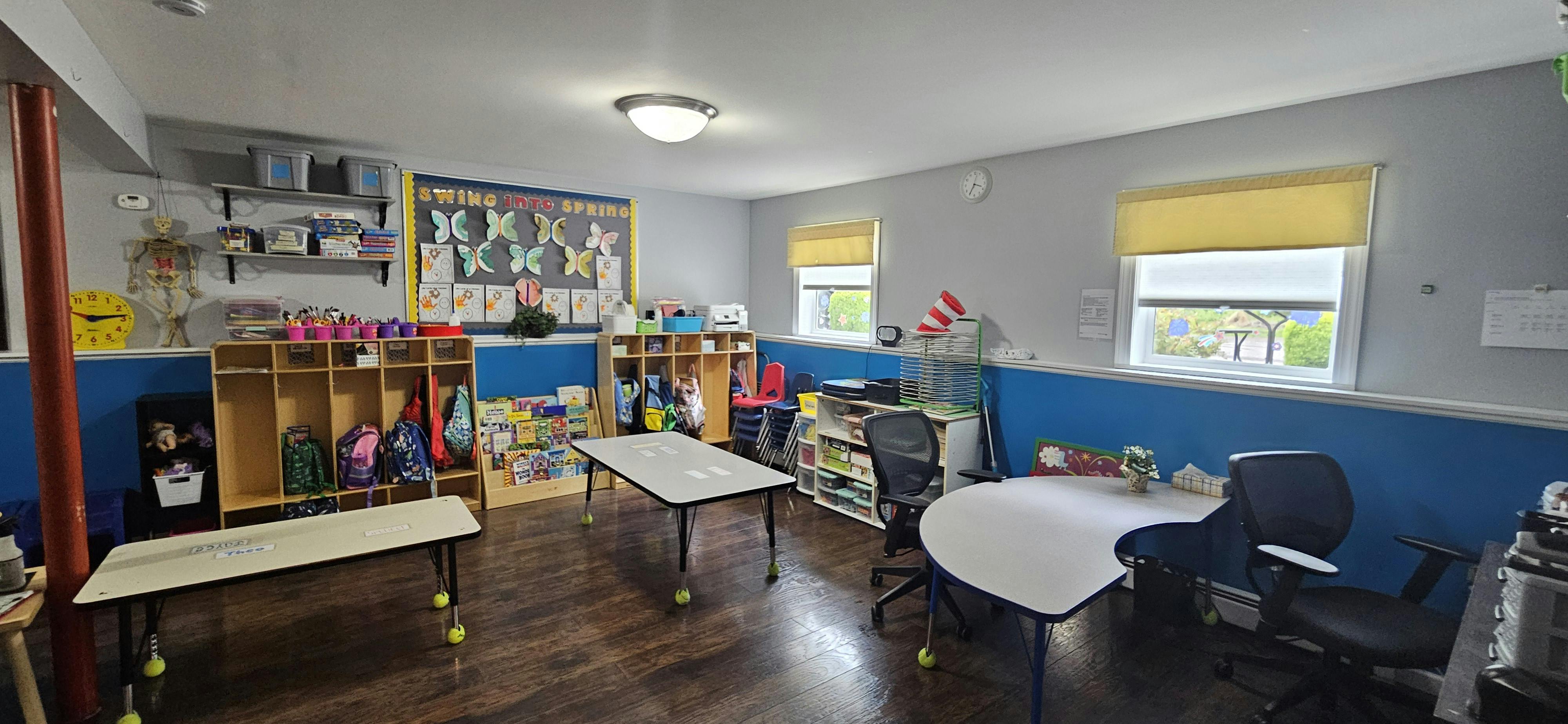 An empty classroom with small tables and chairs, a bulletin board, and storage shelves