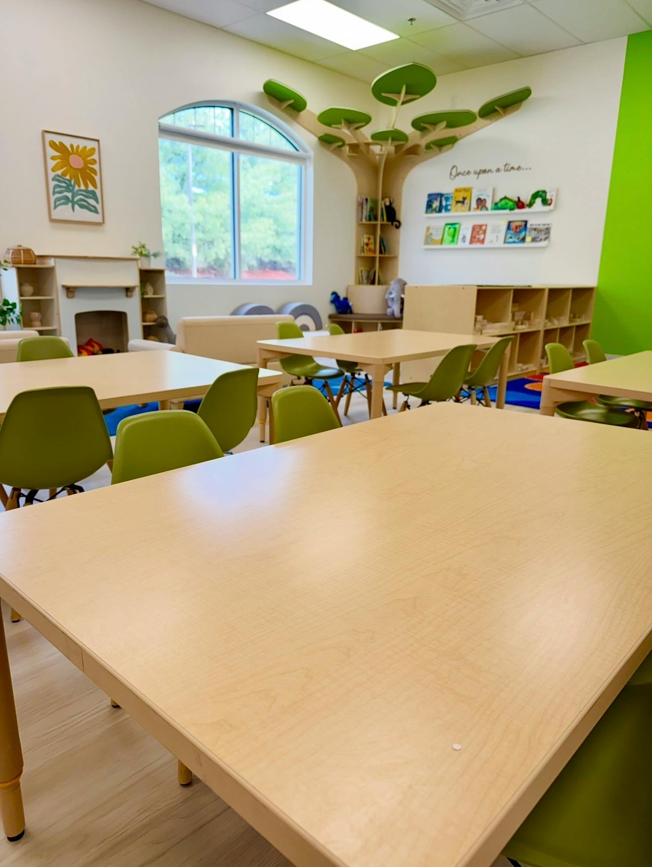 Empty tables and green chairs in a child classroom with a tree wall decoration